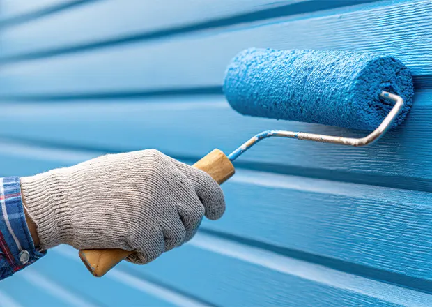 Gloved hand rolling bright blue paint onto house siding